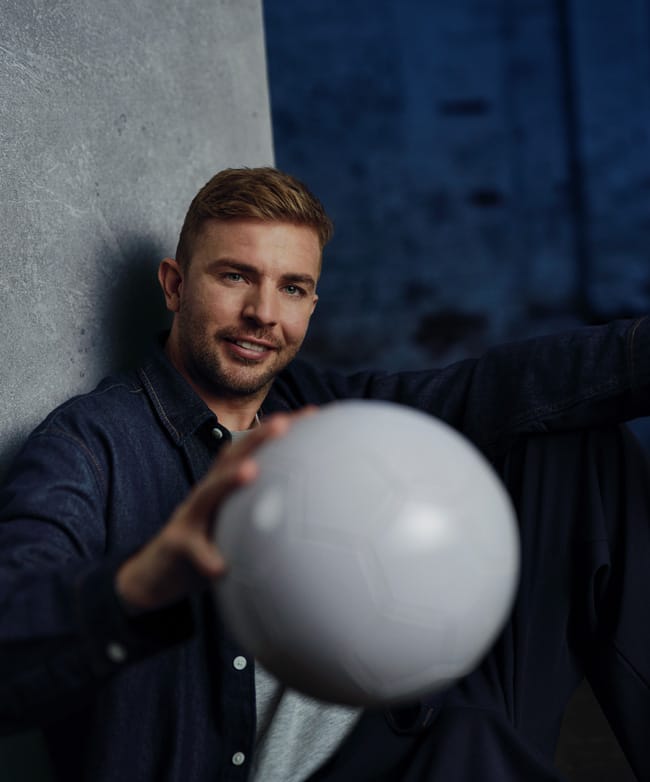 Christoph Kramer lehnt an einer Säule und hält lächelnd einen weißen Fußball in die Kamera.