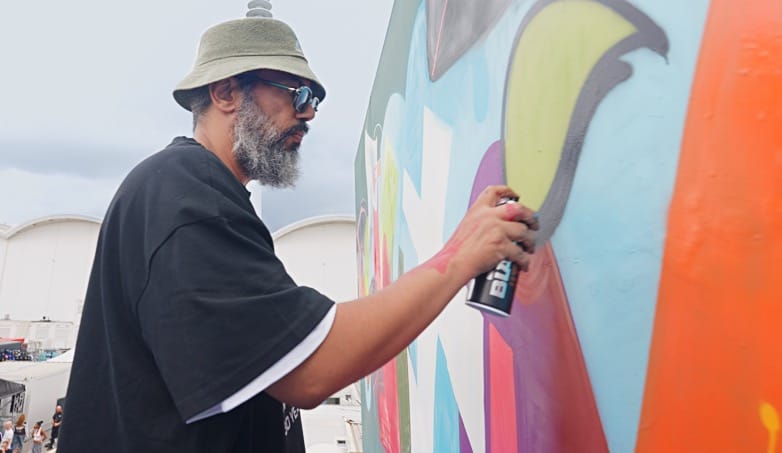 Samy Deluxe mit Sonnenbrille steht an einer Mauer und sprüht mit einer Spraydose ein Graffiti.
