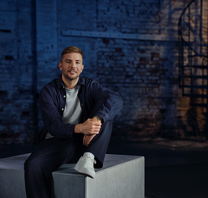 Quadratisches Foto des Fußballweltmeisters und Buchautors Christoph Kramer. Er sitzt im farbigen Interview-Raum mit Industriecharakter auf einem grauen Betonblock.