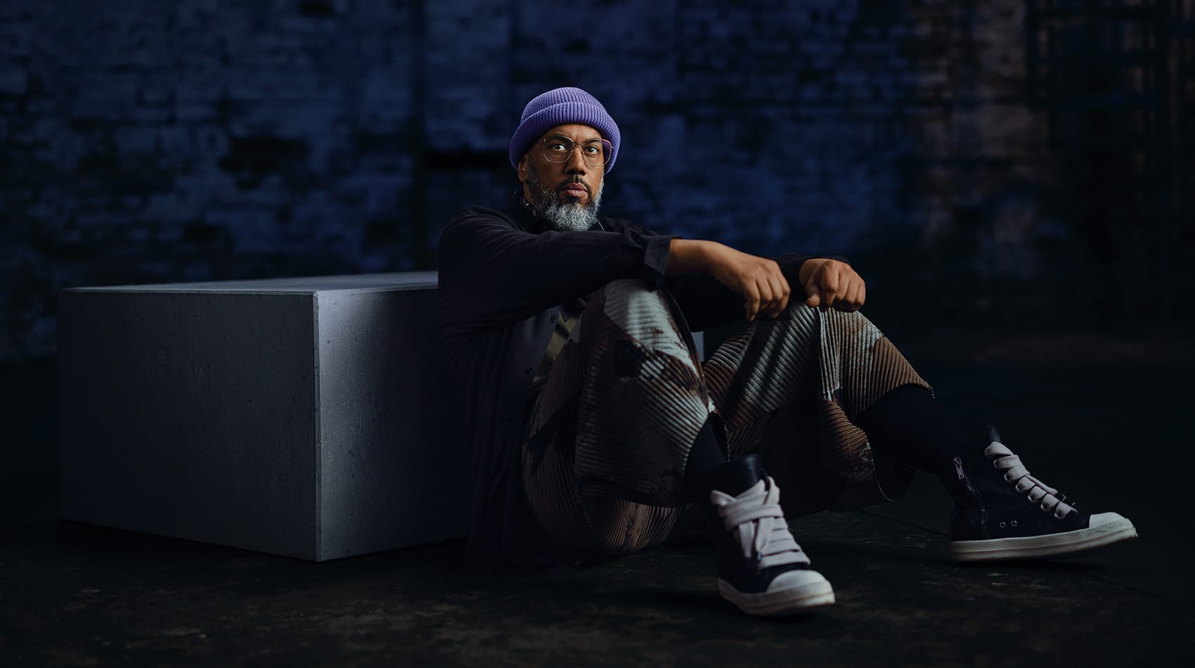 Samy Deluxe sitzt in dunkler Kleidung, gemusterter Hose und lila Beanie am Boden neben einem grauen Betonquader.