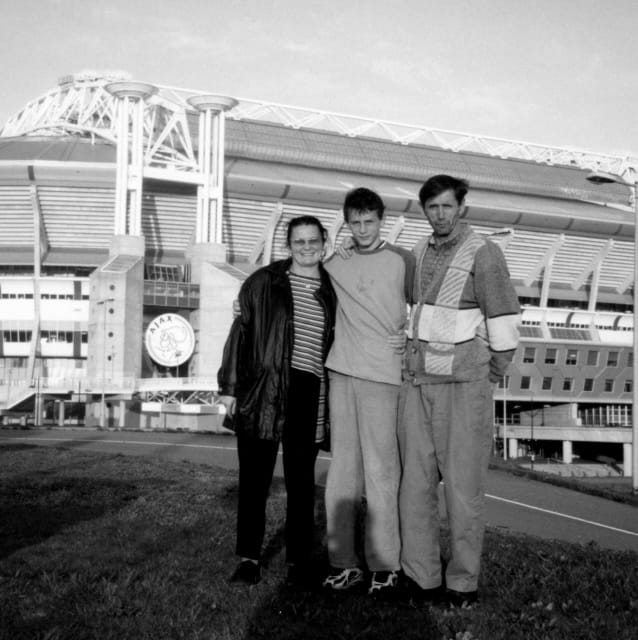 Quadratisches Gruppenfoto des Ex-Fußballprofis Neven Subotić als Teenager mit seinen Eltern in Schwarz-Weiß. Die drei stehen auf dem Rasen vor dem Stadion des Fußballvereins Ajax Amsterdam und blicken stolz direkt in die Kamera.