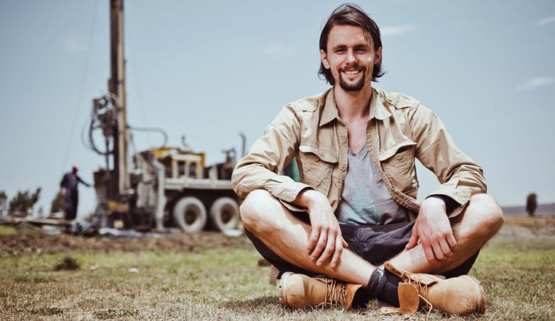 Neven Subotić sitzt im Schneidersitz auf kargem Boden, er lächelt; im Hintergrund eine Bohranlage für Brunnenbau bei Sonne.