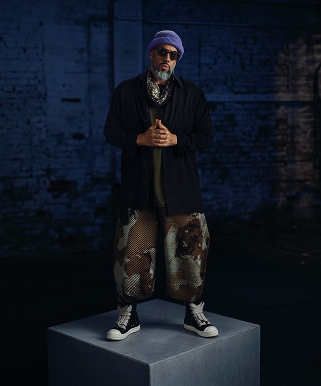 Ganzkörperaufnahme von Samy Deluxe mit Sonnenbrille, der auf einem grauen Betonquader vor blauer Wand steht.