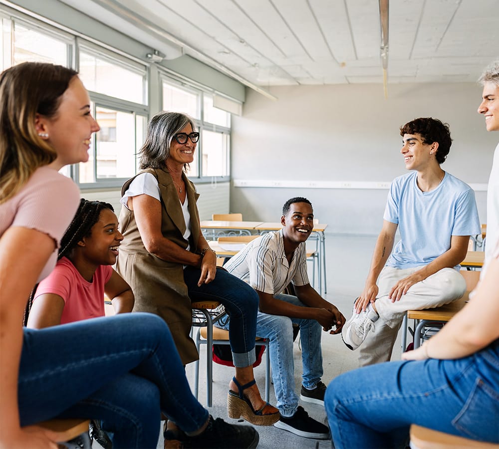 Lehrerin sitzt auf Schultisch und lacht mit Schülern im Klassenzimmer. Offene, fröhliche Gesprächsatmosphäre.