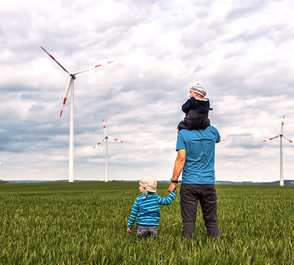 Mann mit zwei Kindern auf einem Feld blickt auf Windkraftanlagen unter bewölktem Himmel.