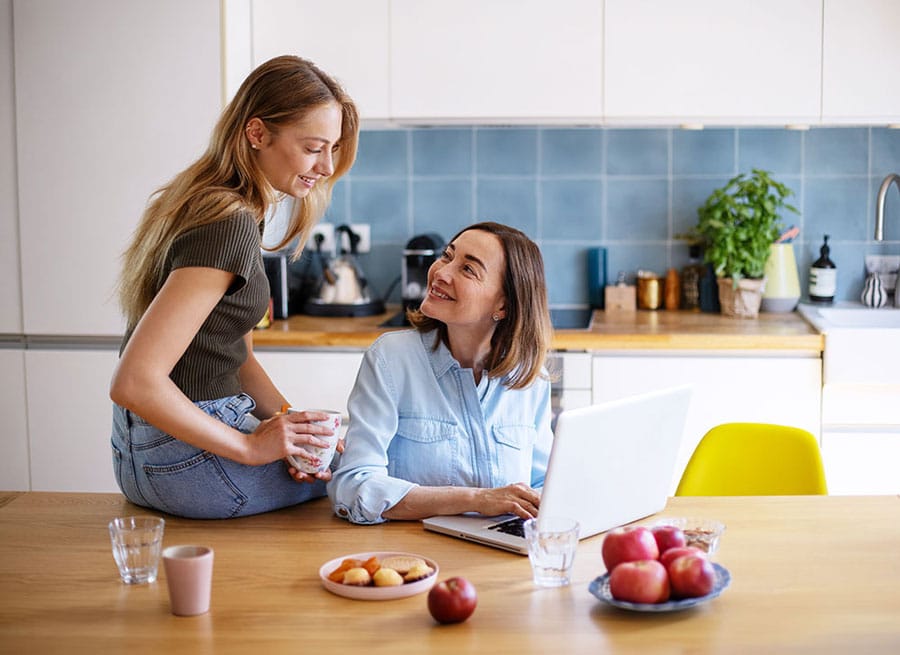 Zwei Frauen lächeln sich in einer Küche vertraut an, eine von ihnen bedient einen Laptop.