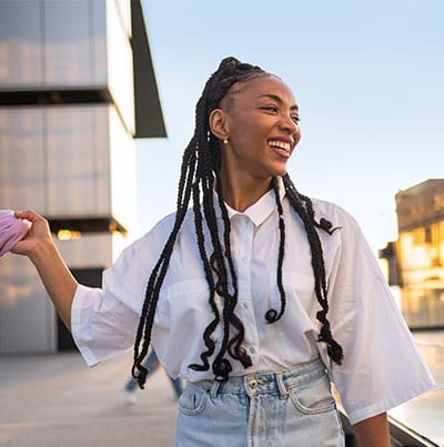 Eine lachende junge Frau mit Braids steht bei Sonnenuntergang vor modernen städtischen Gebäuden.