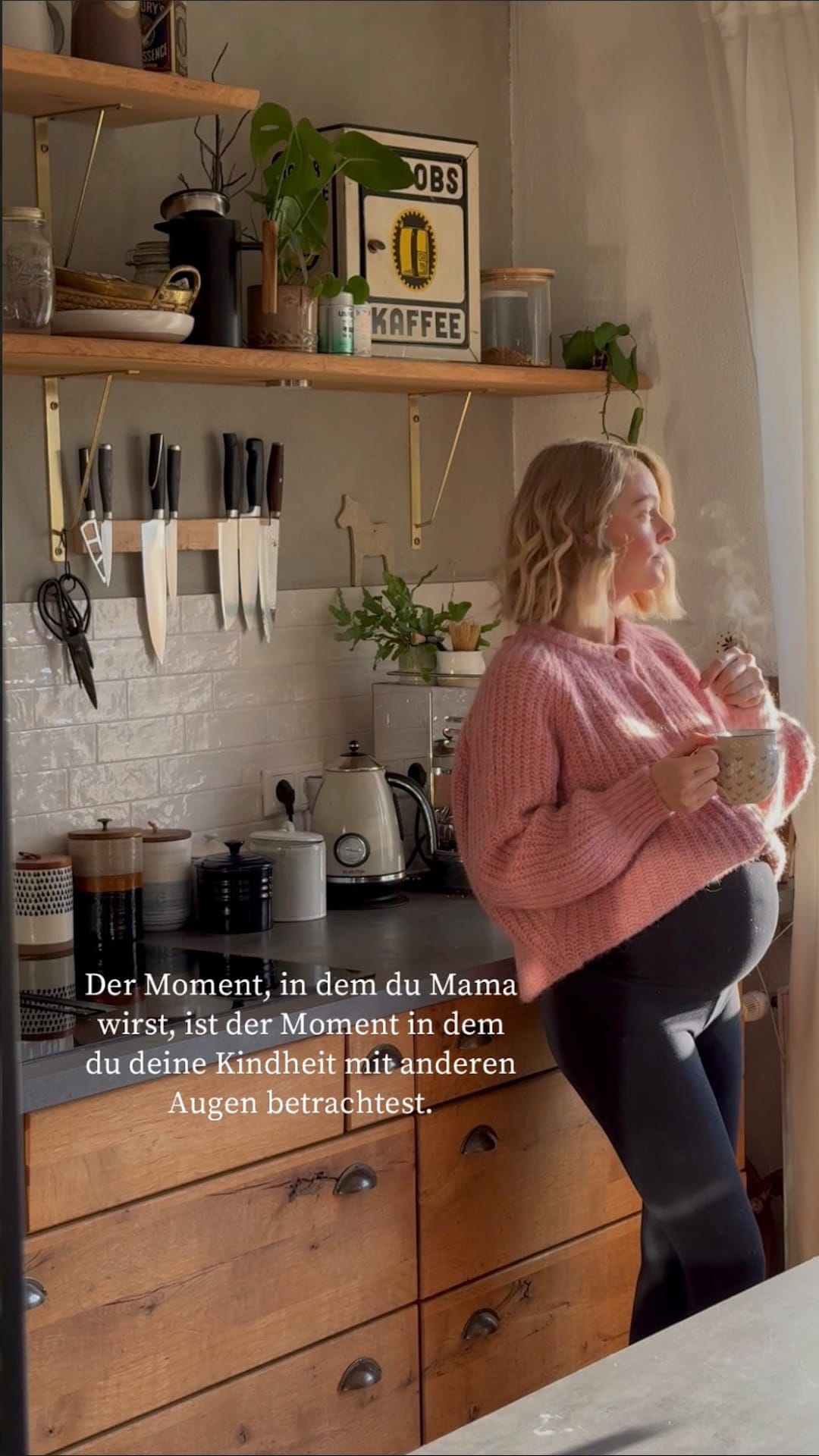Ein vertikales Foto der hochschwangeren Influencerin Larissa (LaryLoves) in einer hellen Küche. Sie trägt einen rosafarbenen Pullover, lehnt sich an der Arbeitsplatte an und blickt auf eine dampfende Tasse, die sie in den Händen hält. Die Atmosphäre ist warm und privat.