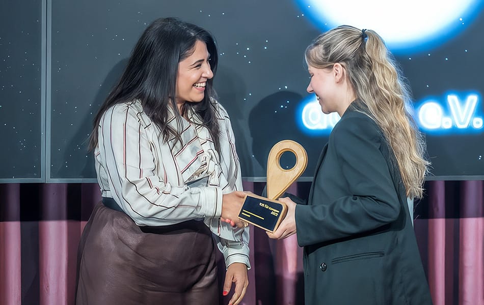 Zwei Frauen schütteln sich freudig die Hand; Übergabe eines hölzernen Awards auf einer Bühne mit Sternen-Hintergrund.