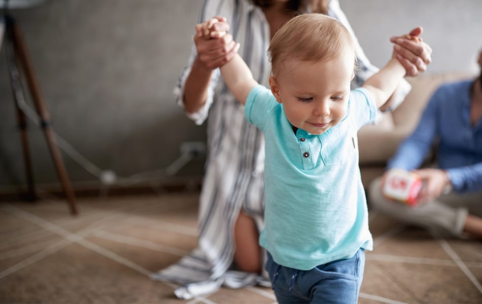 Kleinkind wagt an Mamas Händen erste Schritte; Papa schaut im Hintergrund zu. Familiäre Geborgenheit im Wohnzimmer.