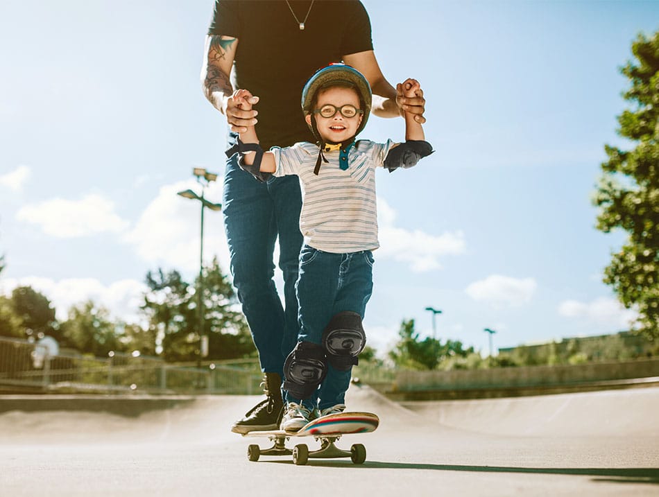 Ein Mann stützt einen lachenden kleinen Jungen mit Helm, der an einem sonnigen Tag Skateboard fährt.