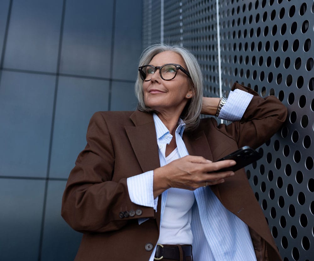 Eine lächelnde ältere Frau mit Smartphone in der Hand lehnt entspannt an einer modernen Hauswand.