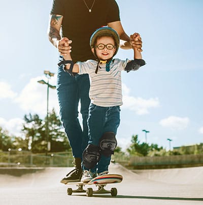 Ein Mann stützt einen lachenden kleinen Jungen mit Helm, der an einem sonnigen Tag Skateboard fährt.